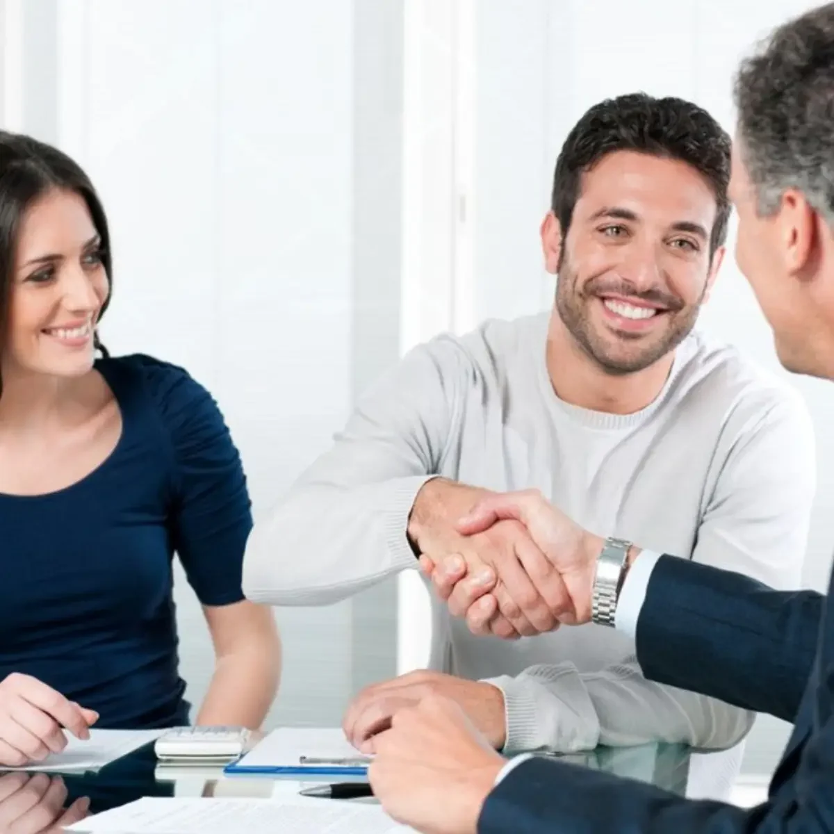 Two people are shaking hands across a glass table, with documents and a calculator visible, suggesting a business agreement.